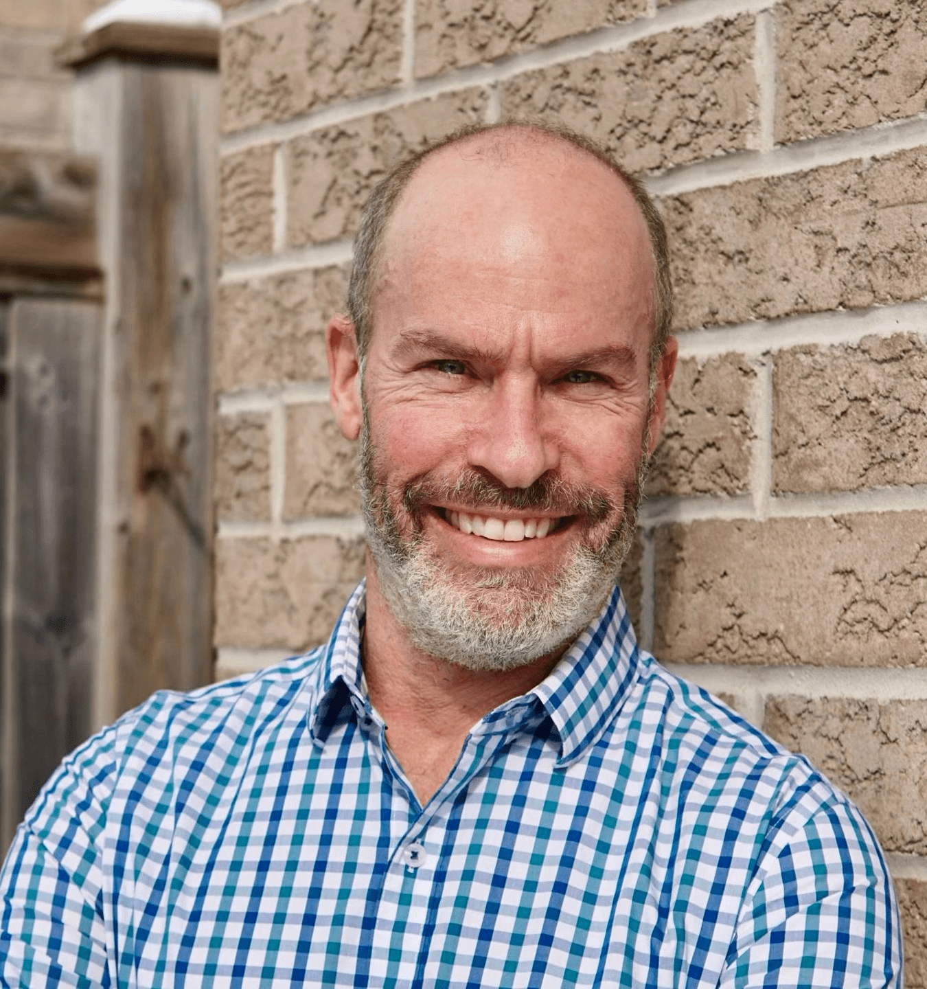 Smiling middle-aged man in a checkered shirt outdoors by a brick wall.