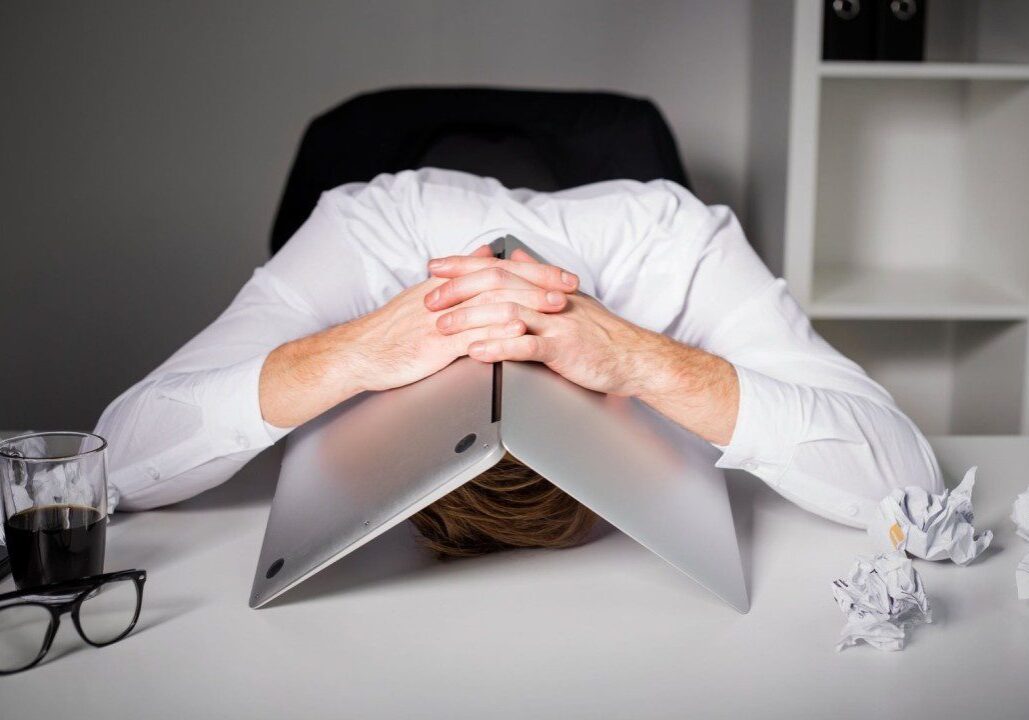 Person overwhelmed, hiding face behind laptop on desk.